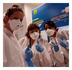 A picture of three researchers in white lab coats and white protective masks all making thumbs up gestures